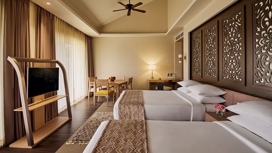 Elegant hotel room with two double beds, carved wooden headboards, ceiling fan, flat-screen TV on a wooden stand, and a seating area near large windows with sheer curtains - Voyage The Solitaire Bliss.
