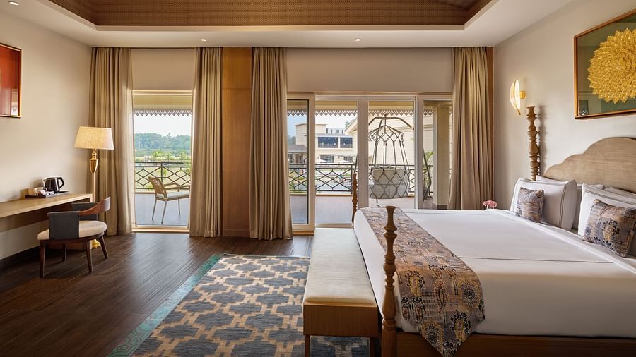 A well-lit hotel room featuring a king size bed with a patterned throw, a wooden desk and chair, and a balcony with a swing offering a view of a cityscape. The room has a wooden ceiling and a patterned rug - Voyage The Solitaire Bliss.
