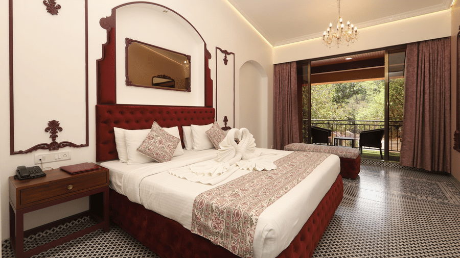 Moghul Room at Zaras Resort with elegant décor, red accents, swan towel art on bed, balcony view of greenery, and ambient lighting.