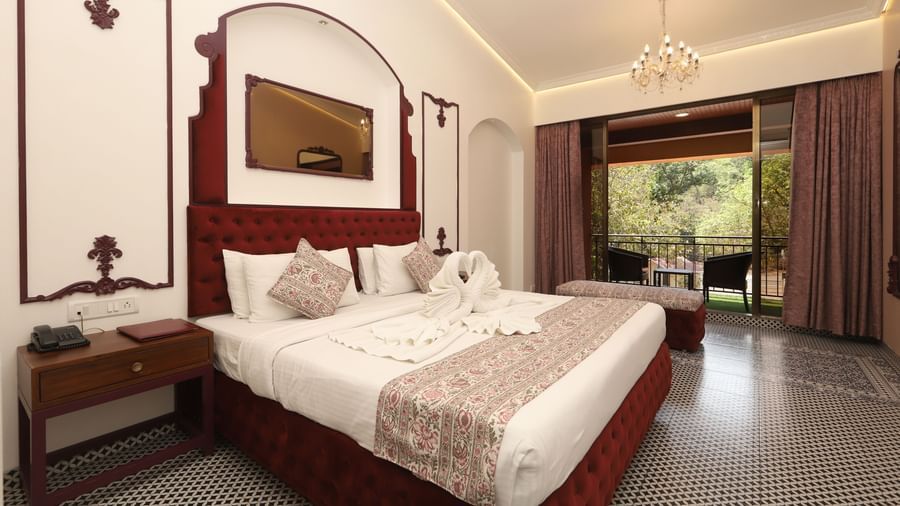 Moghul Room at Zaras Resort with elegant décor, red accents, swan towel art on bed, balcony view of greenery, and ambient lighting.