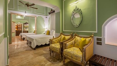 A charming suite at Dera Rawatsar, Jaipur, features two comfortable beds, stylish armchairs, an ornate mirror, and elegant archway details.
