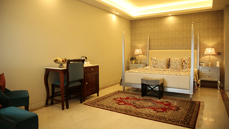 A well-lit bedroom with a four-poster bed, a desk with a chair, a patterned rug, and soft lighting from a recessed ceiling, creating a warm atmosphere | Dileep Kothi, Jaipur