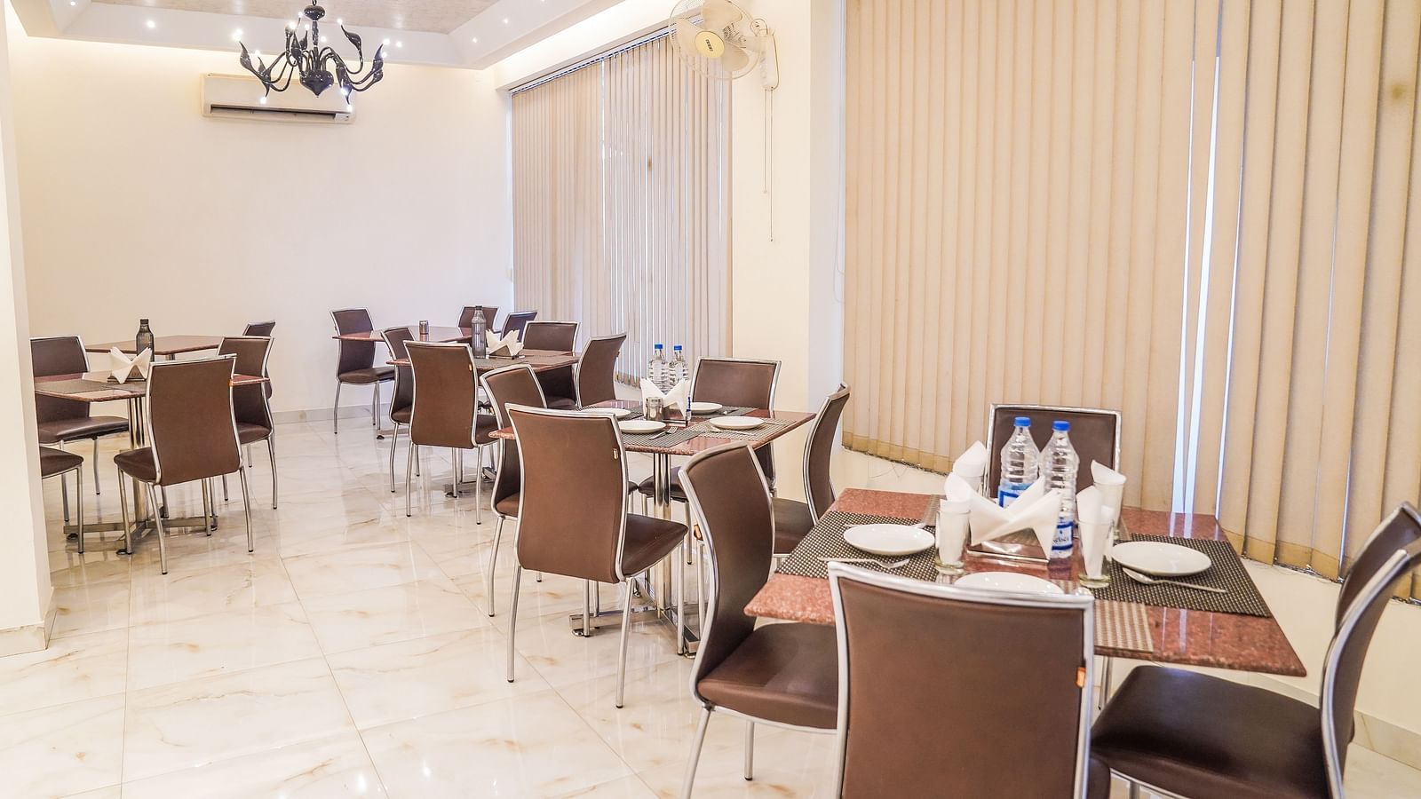 An elegant dining area with multiple tables and chairs is illuminated by a modern chandelier and wall lights - Asian Suites Cyber Park, Gurgaon