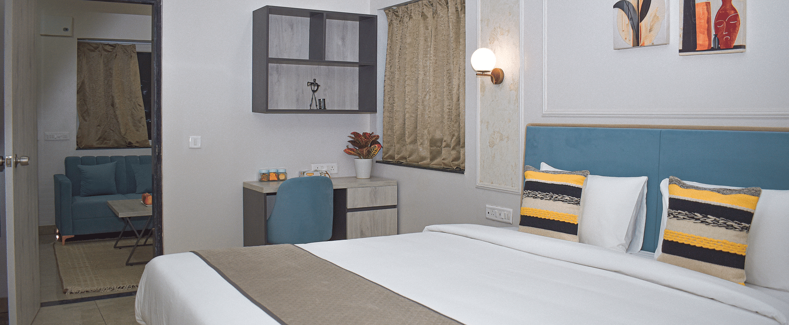 A hotel bedroom with a bed, a desk and chair, a wall cabinet, a window with curtains, and a doorway leading to a living area with a blue sofa - Urban Suites by BluSalzz, Jaipur