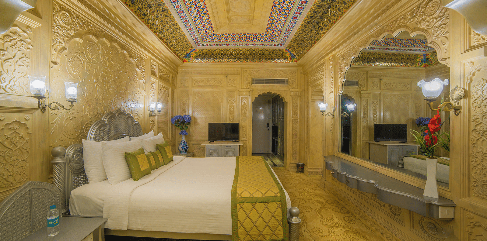 A palatial room with exquisite carvings, muted tones, and plush bedding for a regal experience A grand hotel suite with an opulent red and white decor, large bed, and seating area, reflecting a traditional style at Sitara Luxury Hotel in Hyderabad near Ram