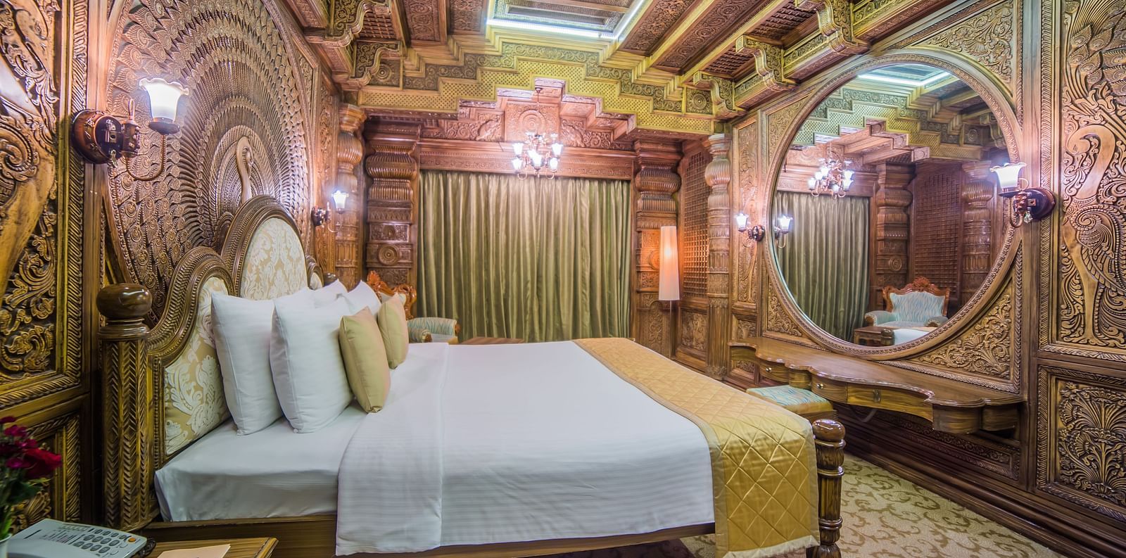 An intricately carved room with traditional motifs, a plush bed, and rich wooden decor A grand hotel suite with an opulent red and white decor, large bed, and seating area, reflecting a traditional style at Sitara Luxury Hotel in Hyderabad near Ramoji Film