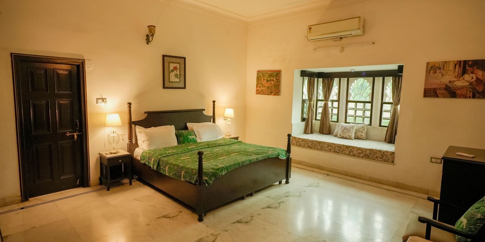 A wide shot of the heritage room at Mahendra Niwas, featuring a large, dark wooden bed with a vibrant green patterned duvet, and a cozy window seat.
