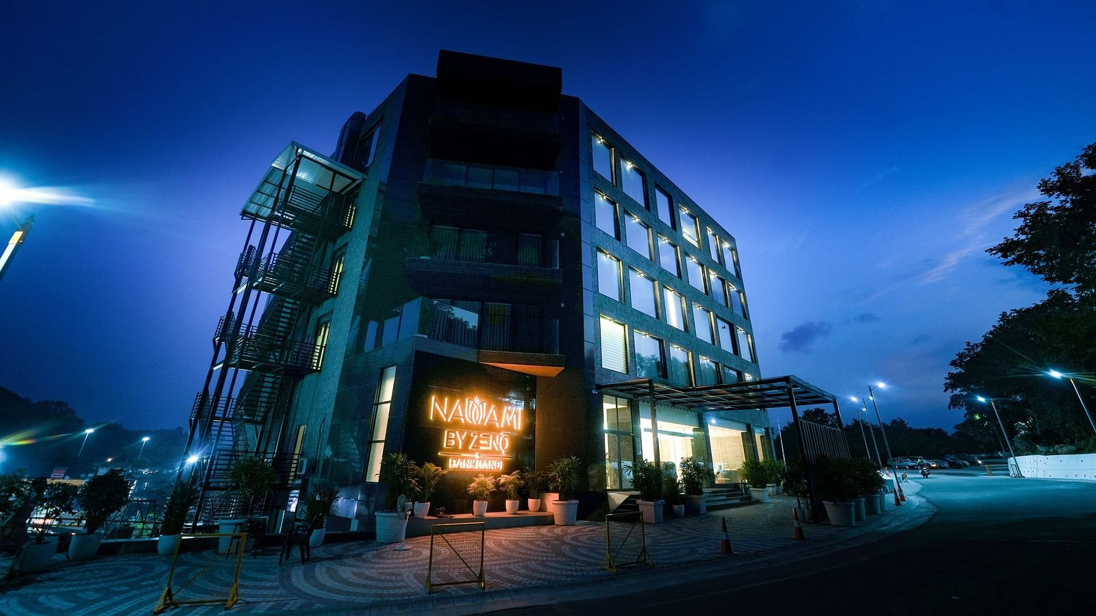 A hotel building set against a dark sky, illuminated by the brightly lit signage displaying the name of the hotel - NAMAMI BY ZENQ BANKHANDI