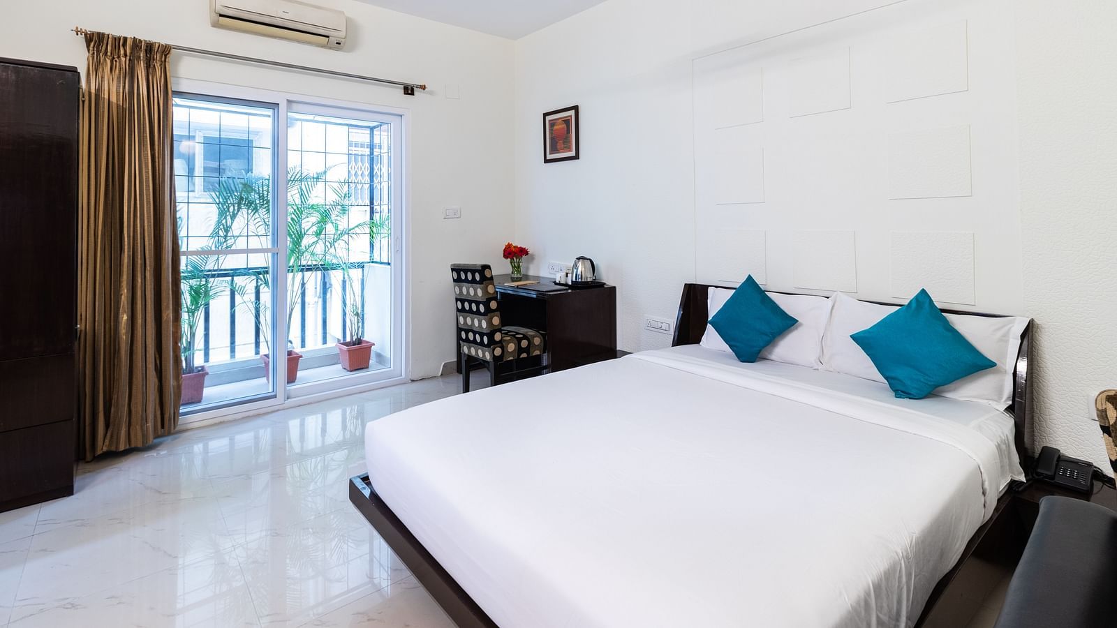 A room with a double bed with blue pillows, a work desk and chair, and glass door leading to the balcony at Sanctum Suites Domlur, Bangalore.