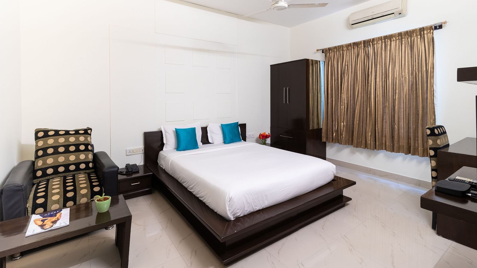 A tidy bedroom with a double bed, a sofa, and a white ceiling fan at Sanctum Suites Domlur, Bangalore.