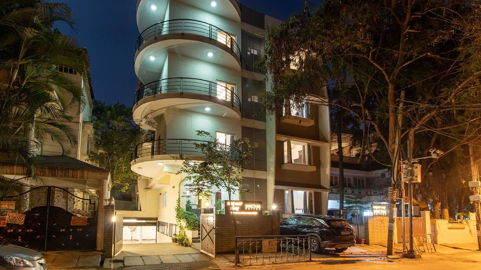 An exterior view of Sanctum Suites Indiranagar, Bangalore showcasing the cylindrical building illuminated at night along with greenery around.