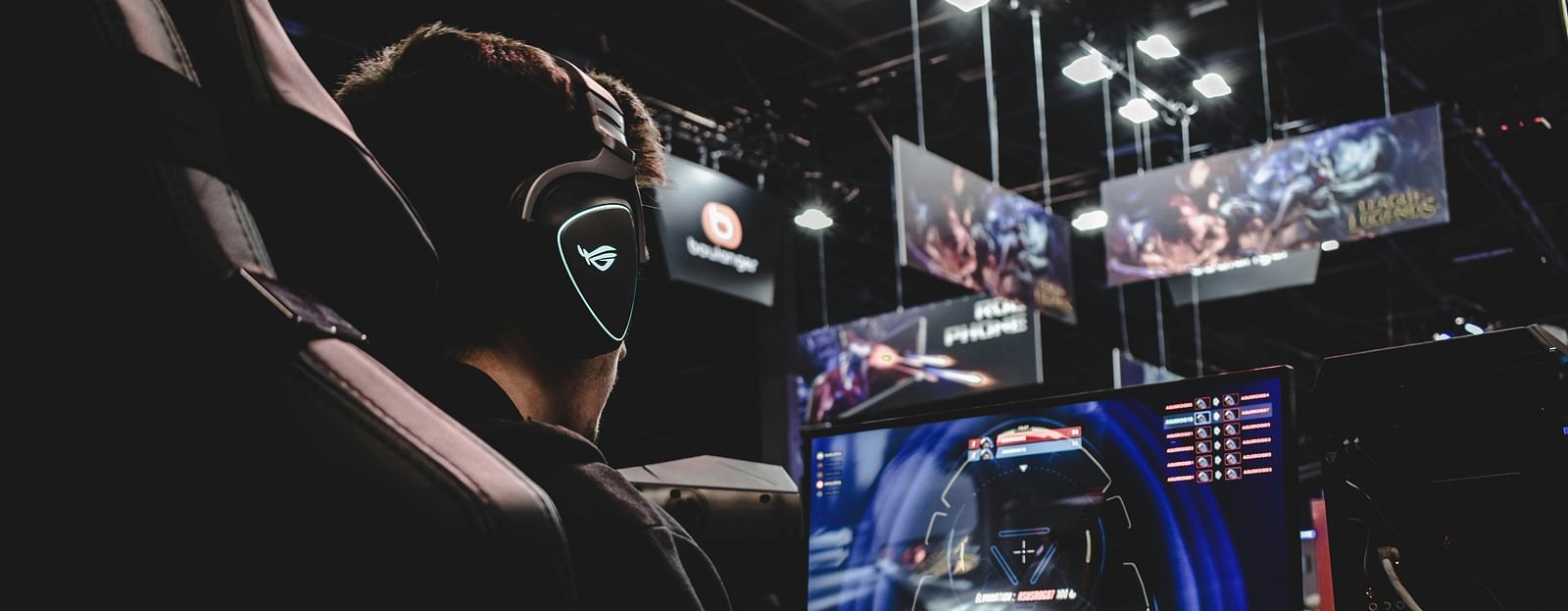Side view from the back of a man wearing gaming equipment and playing games on a machine. There are screens in the background.