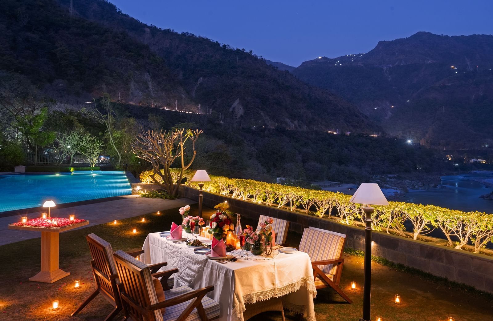A dinner setup by a pool with mountains in the background. The table is adorned with candles and flowers.