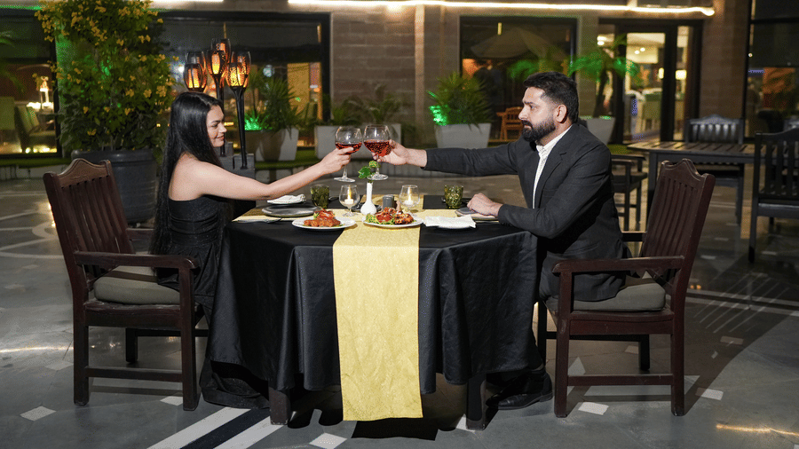 A couple sharing a meal at an outdoor dining setup at Park Plaza Ludhiana with dark rattan chairs black tablecloth warm ambient lighting and green foliage in the background.