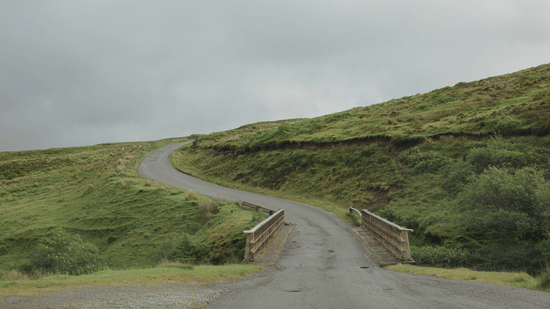 A winding road passing through green hills under a cloudy sky.