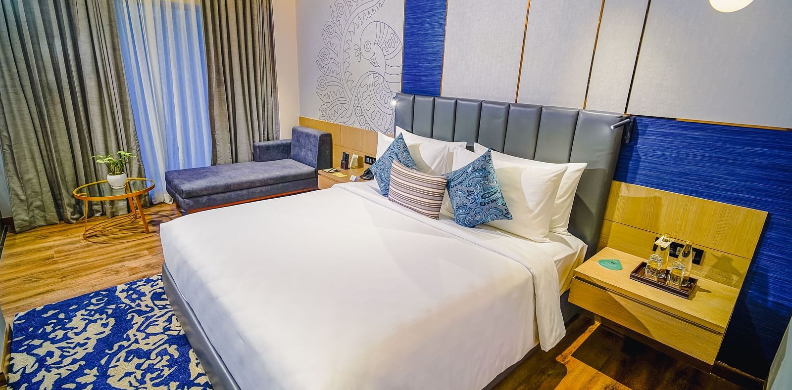 An image of a hotel room in Madurai with a large bed, a blue carpet, a couch and a night stand