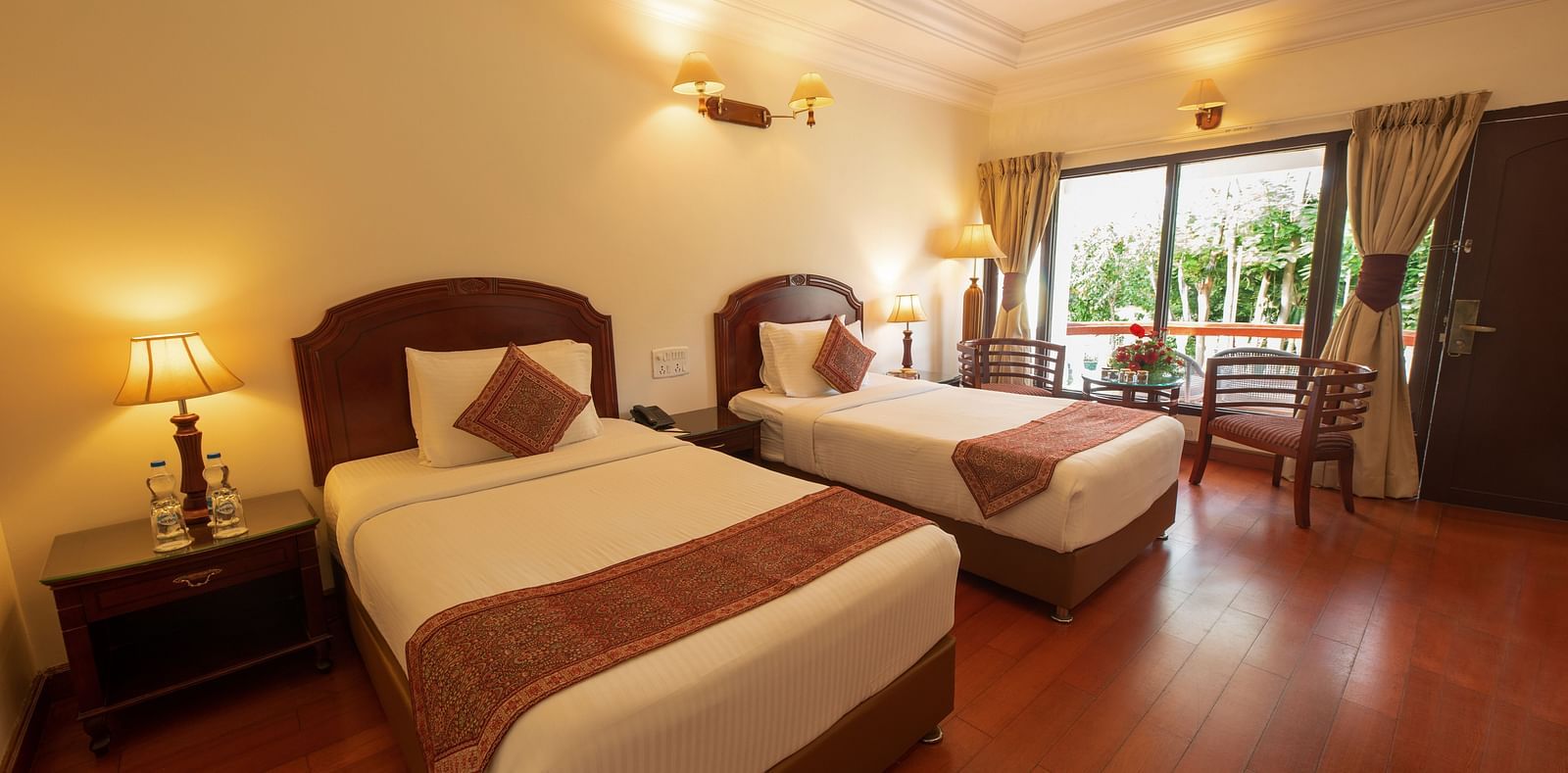 An image of a River View Room with balcony featuring two single beds, bed side table, lamps, ceiling fan, and an expansive glass door leading to the balcony - GReaT Trails River View Resort Thanjavur By GRT Hotels