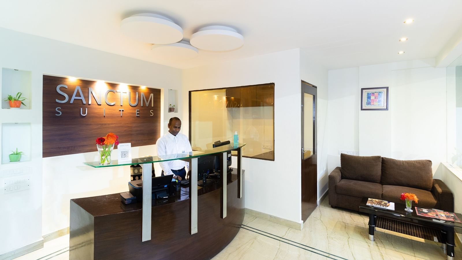 The reception area at Sanctum Suites Domlur, Bangalore featuring a reception desk, a brown sofa, and a coffee table.