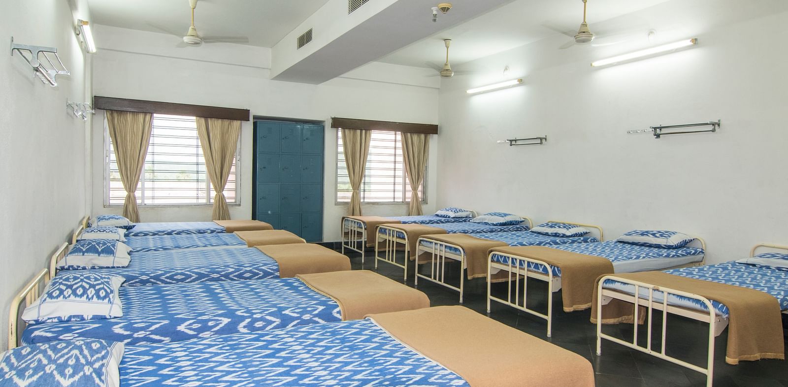 Beds with bedding are arranged in two rows in a room with two windows at Hotel Sahara - Comfortable Dormitory in Ramoji Film City, Hyderabad.