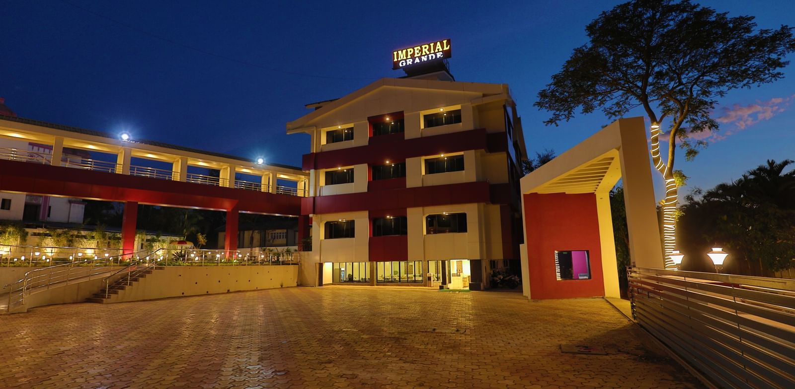 The exterior of Imperial Grande at night, showing a multi-story building with illuminated logo and a paved courtyard reflecting the lights.