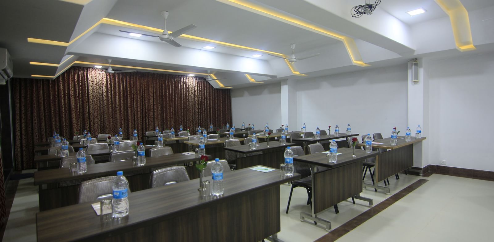 A well-lit conference room at Krushnai Resort with long rectangular tables arranged in class room style with chairs around them and a water bottle on each table.