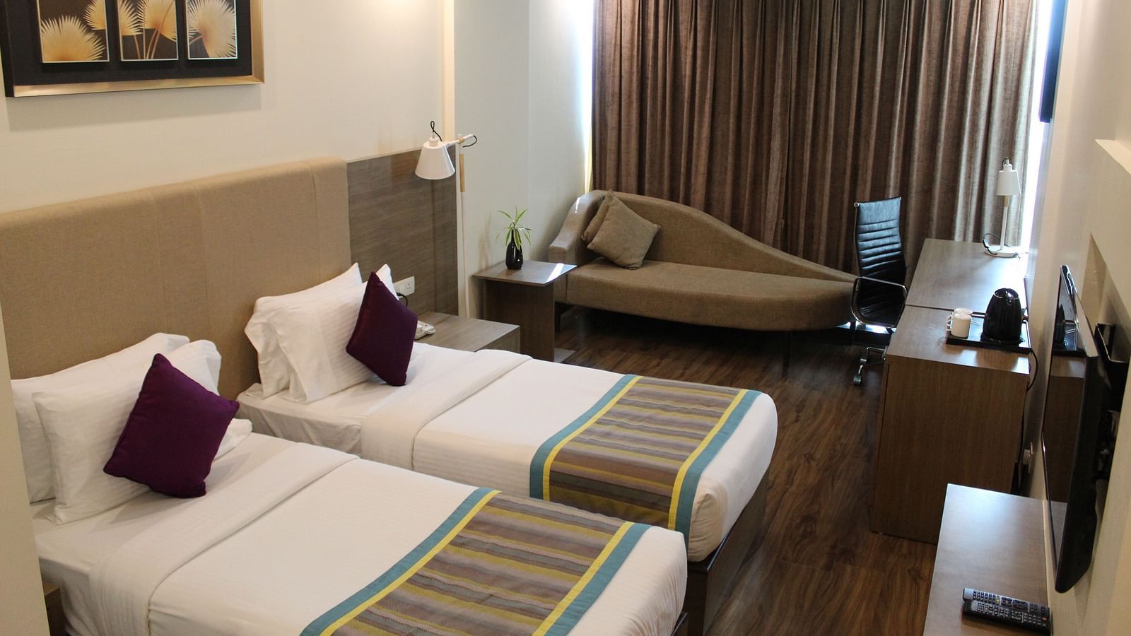 An image showing a pair of twin beds with lush decors, a long sofa with cushion, and a flat screen TV, inside a room with closed curtains.- Golden Suites, Gurgaon