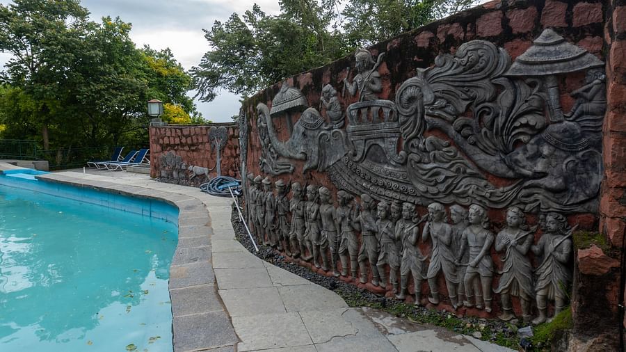 A swimming pool at Mango Hill, Yercaud, with a long, brown wall featuring a detailed bas-relief sculpture depicting numerous figures.