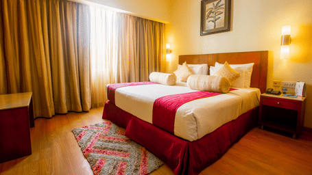 Hotel room at Featherlite Evoma Hotel with a bed dressed in white and maroon linen, wooden furnishings and long curtains by the window nearby.