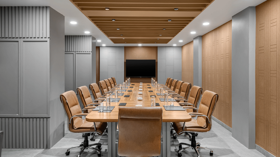 A formal conference room featuring a long, light wood meeting table surrounded by brown upholstered chairs, with grey panelled walls and a wood-panelled ceiling | Voyage Eastin Suites