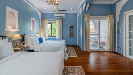 A blue-walled bedroom with two twin beds, hardwood floors, a chandelier, and French doors leading to a balcony | Dera Rawatsar, Jaipur