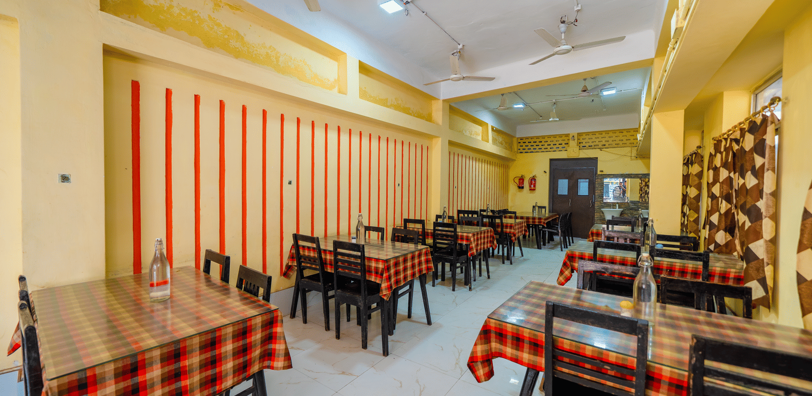 A brightly-lit, empty hotel dining hall with small tables covered in red and black checkered tablecloths and matching chairs - Hotel Dolphin Puri