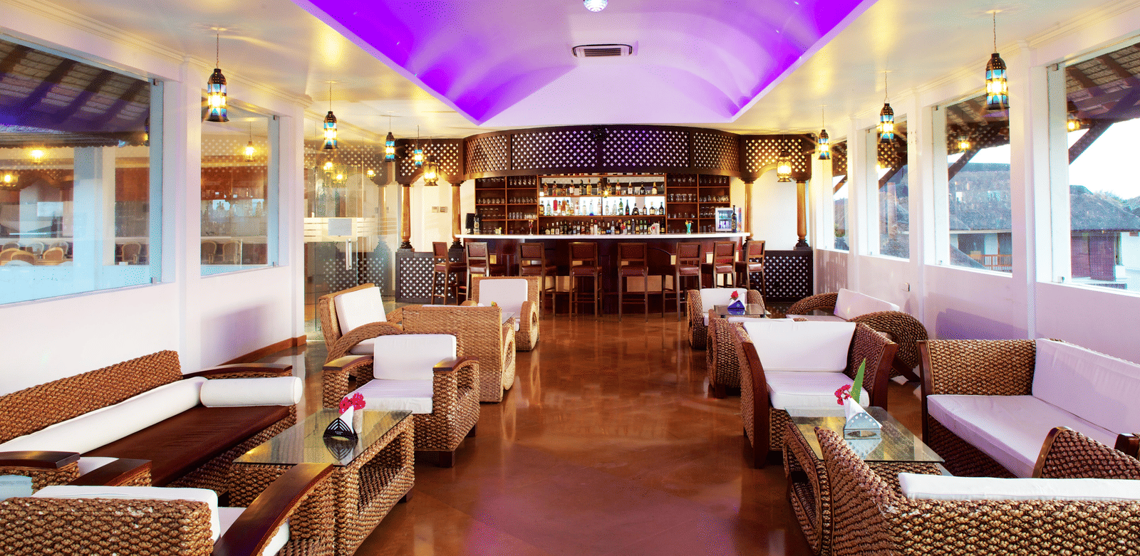 An indoor lounge bar area with wicker sofas, white cushions, and a curved bar counter under a purple-lit ceiling | Le Pondy
