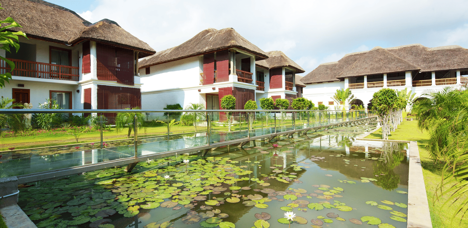A transparent glass bridge with metal railings built over a pond covered with lily pads next to 2-storey resort buildings | Le Pondy