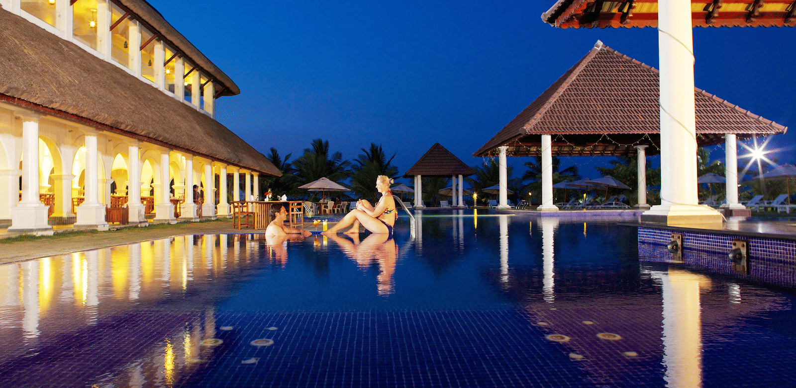 An outdoor swimming pool with blue tiles illuminated at night next to lit resort buildings and pavilions | Le Pondy