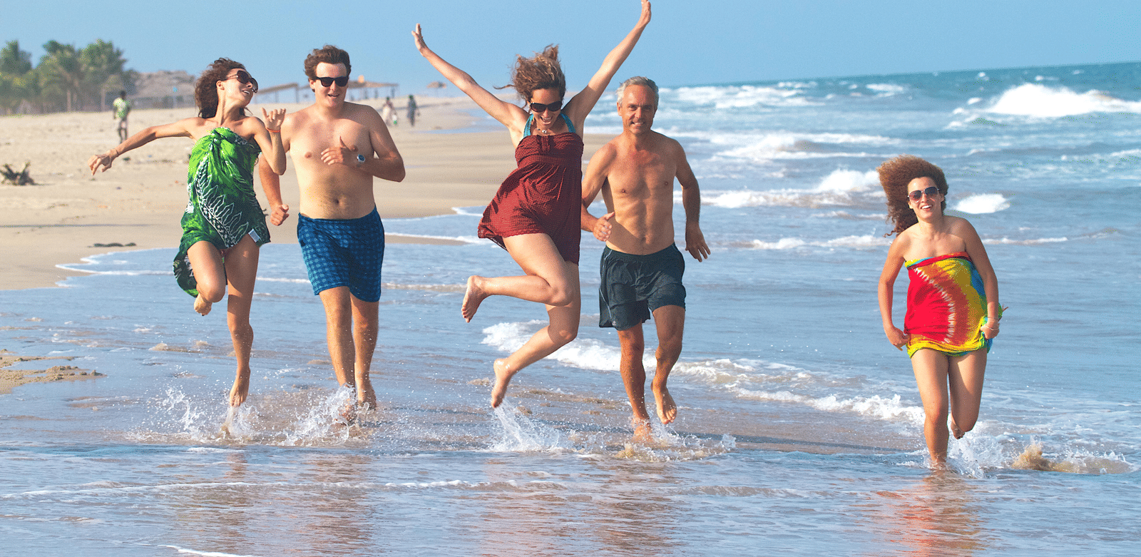 A group of 5 people running and jumping playfully along the shoreline of a sandy beach | Le Pondy