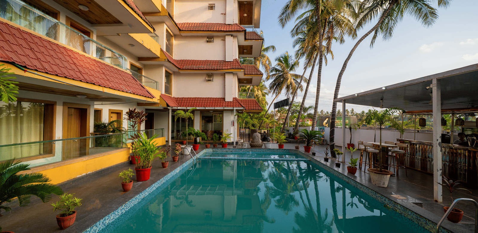 A sunny outdoor swimming pool is located within the courtyard surrounded by palm trees and buildings at Perfectstayz Premium Goa Continental.