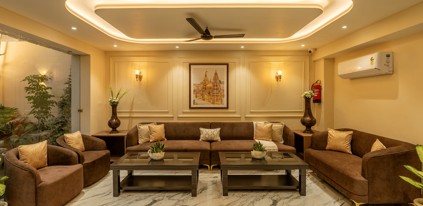 Front view of the lobby lounge at Perfectstayz Premium Lavender, Varanasi with 2 brown sofas, 2 armchairs, 2 coffee tables, cove lighting, and a framed temple print.