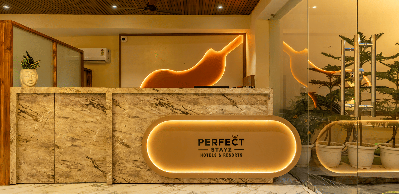 Marble reception counter at Perfectstayz Premium Lavender, Varanasi with a backlit brand logo, a glowing bird-shaped wall panel, potted plants, and a glass door.