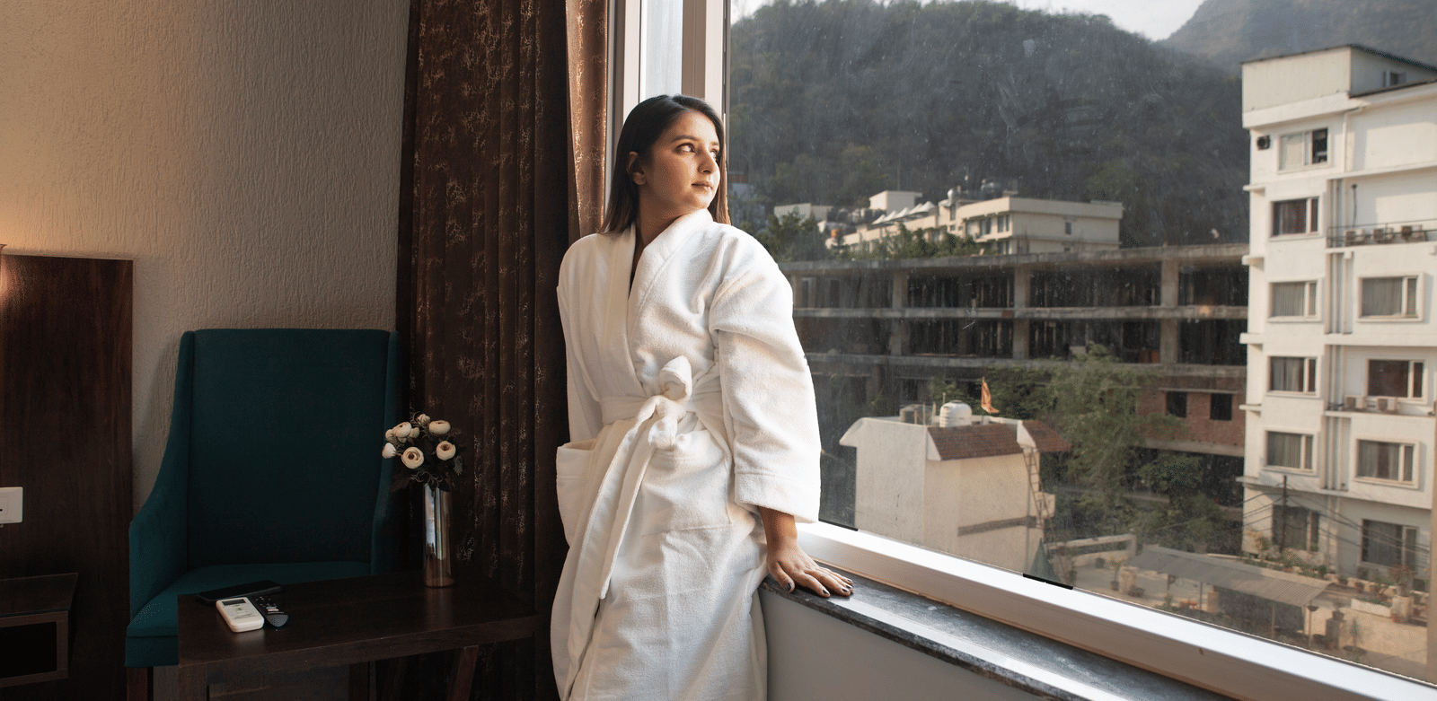 A woman in a bathrobe in a bedroom at Perfectstayz Premium Laxmi Heritage stands looking out a window at mountains and buildings.