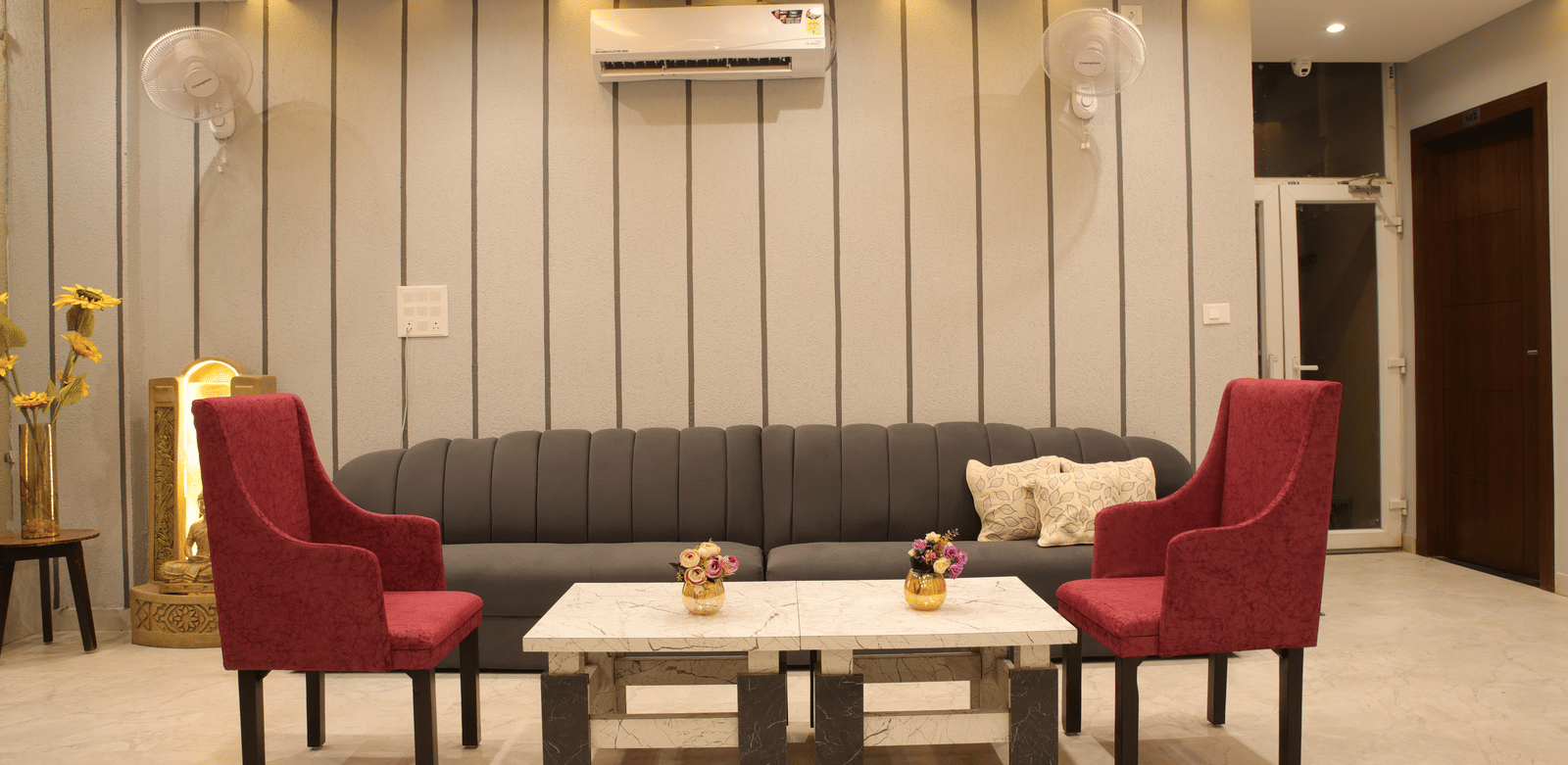 A lobby area at Perfectstayz Premium Laxmi Heritage with a sofa, 2 armchairs, 2 coffee tables, an A/C, and striped wall panelling with decorative lighting.