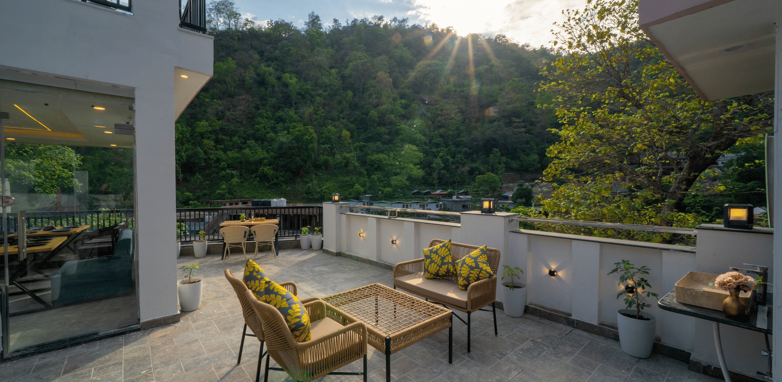 An outdoor terrace with tables, wicker chairs, cushions, and a view of hills under sunlight in Perfectstayz Premium Mystic Falls, Rishikesh.