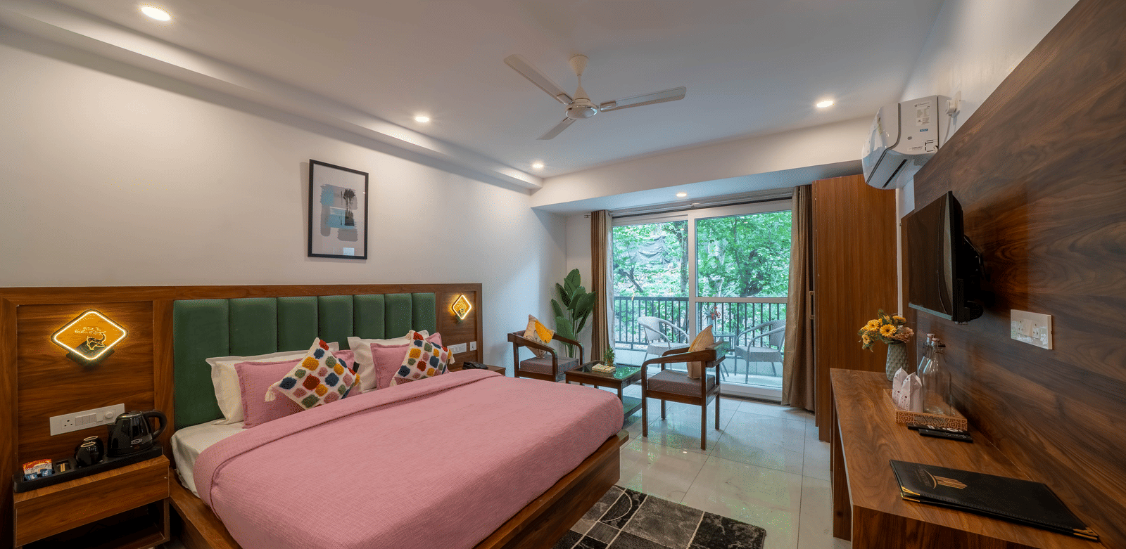 A room with a bed, a headboard, a television, a seating area with 2 chairs and a table, and a glass door leading to a balcony in Perfectstayz Premium Mystic Falls, Rishikesh.