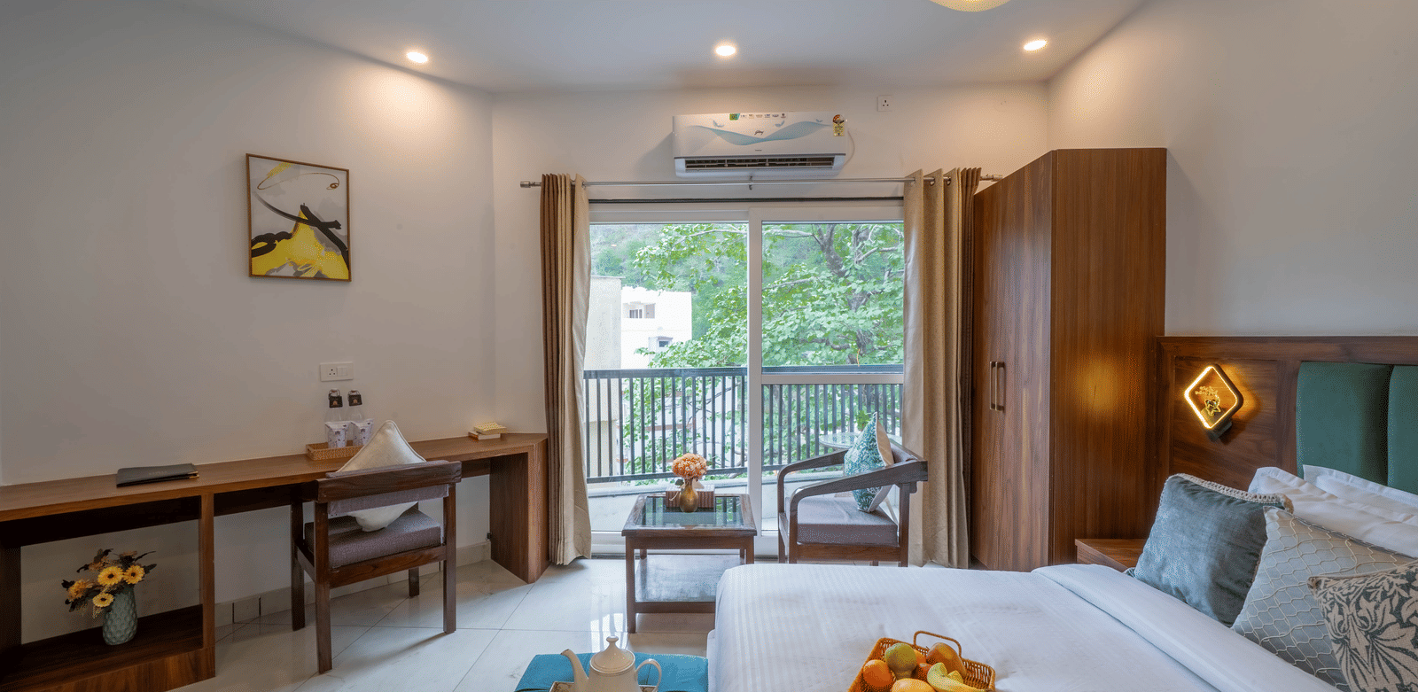 A room with a bed, a headboard, a wooden desk with a chair, a wardrobe, and a tray of fruit on the bed in Perfectstayz Premium Mystic Falls, Rishikesh.