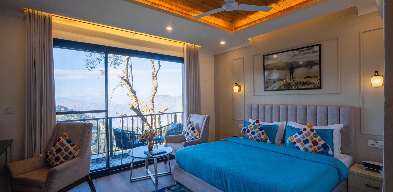 A wide-angle shot of the Executive Room With Balcony & Valley View featuring a double bed with blue sheets and 2 chairs overlooking the valley from the balcony at Perfectstayz Premium Westend Mall Road, Mussoorie.