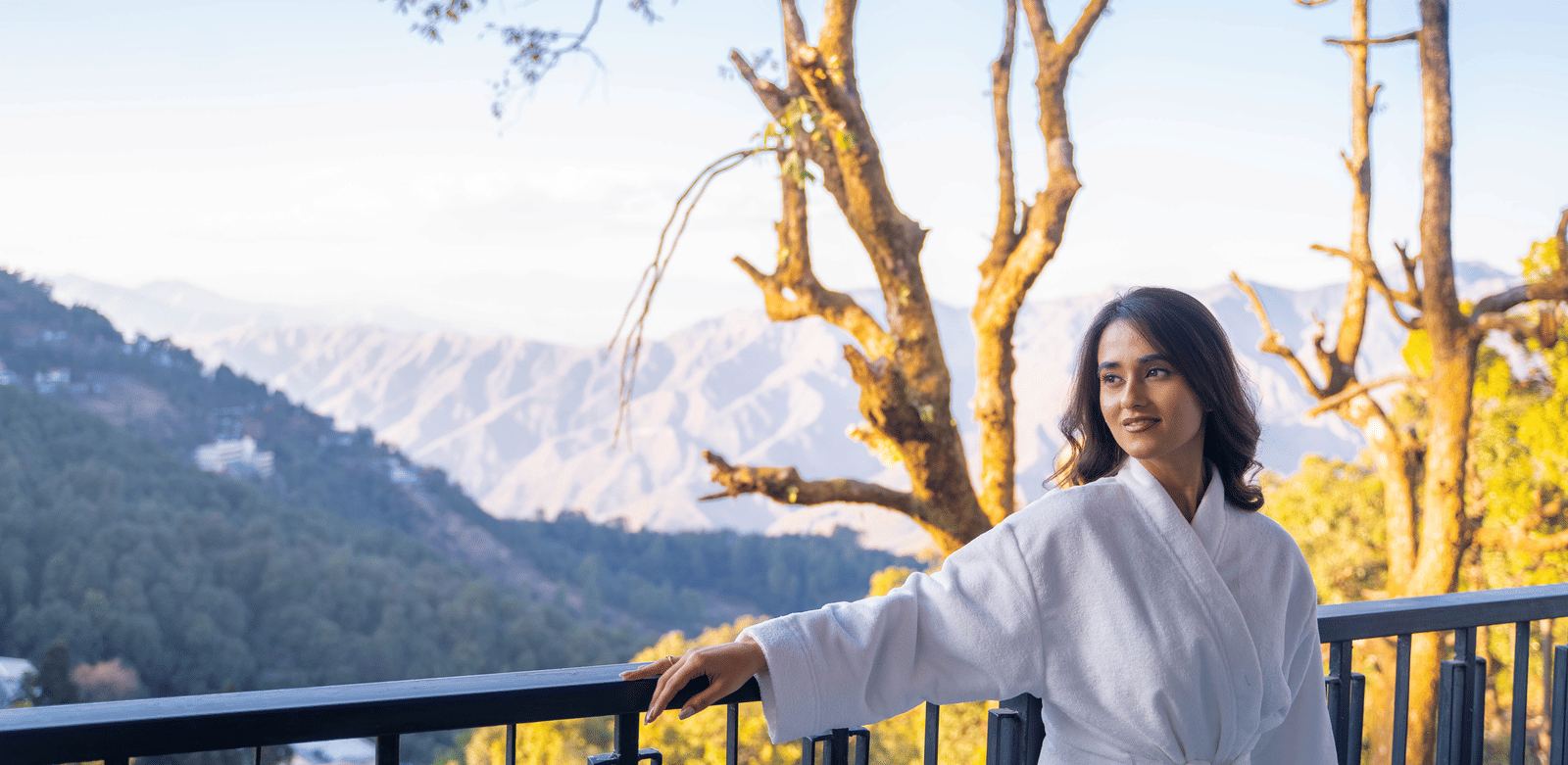 A woman dressed in a bathrobe smiling at the balcony of Perfectstayz Premium Westend Mall Road, Mussoorie, with beautiful mountain views in the background.