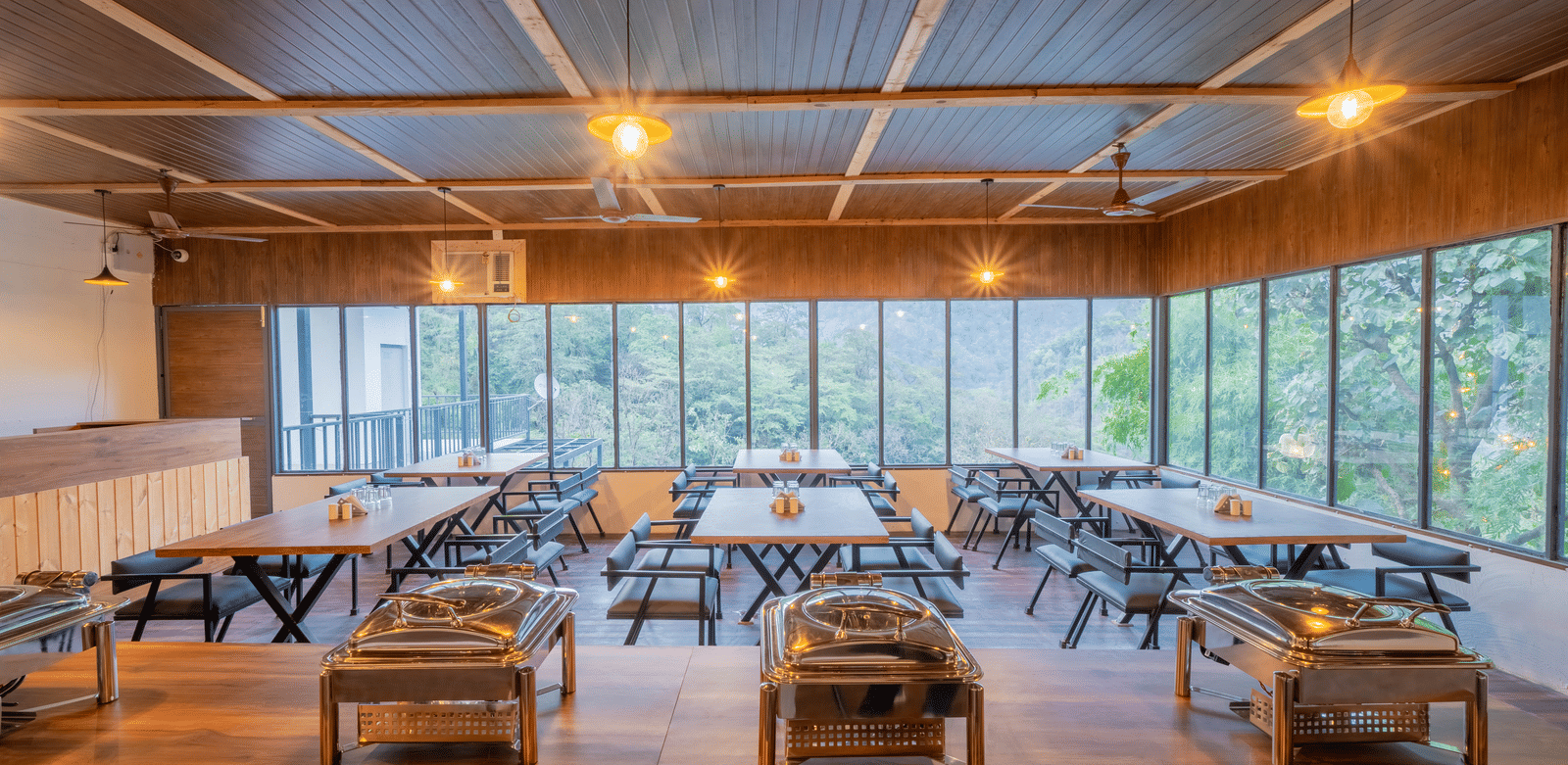 The dining hall at Perfect Stayz The Jungle Resort has long tables, chairs, chafing dishes, and windows overlooking trees.