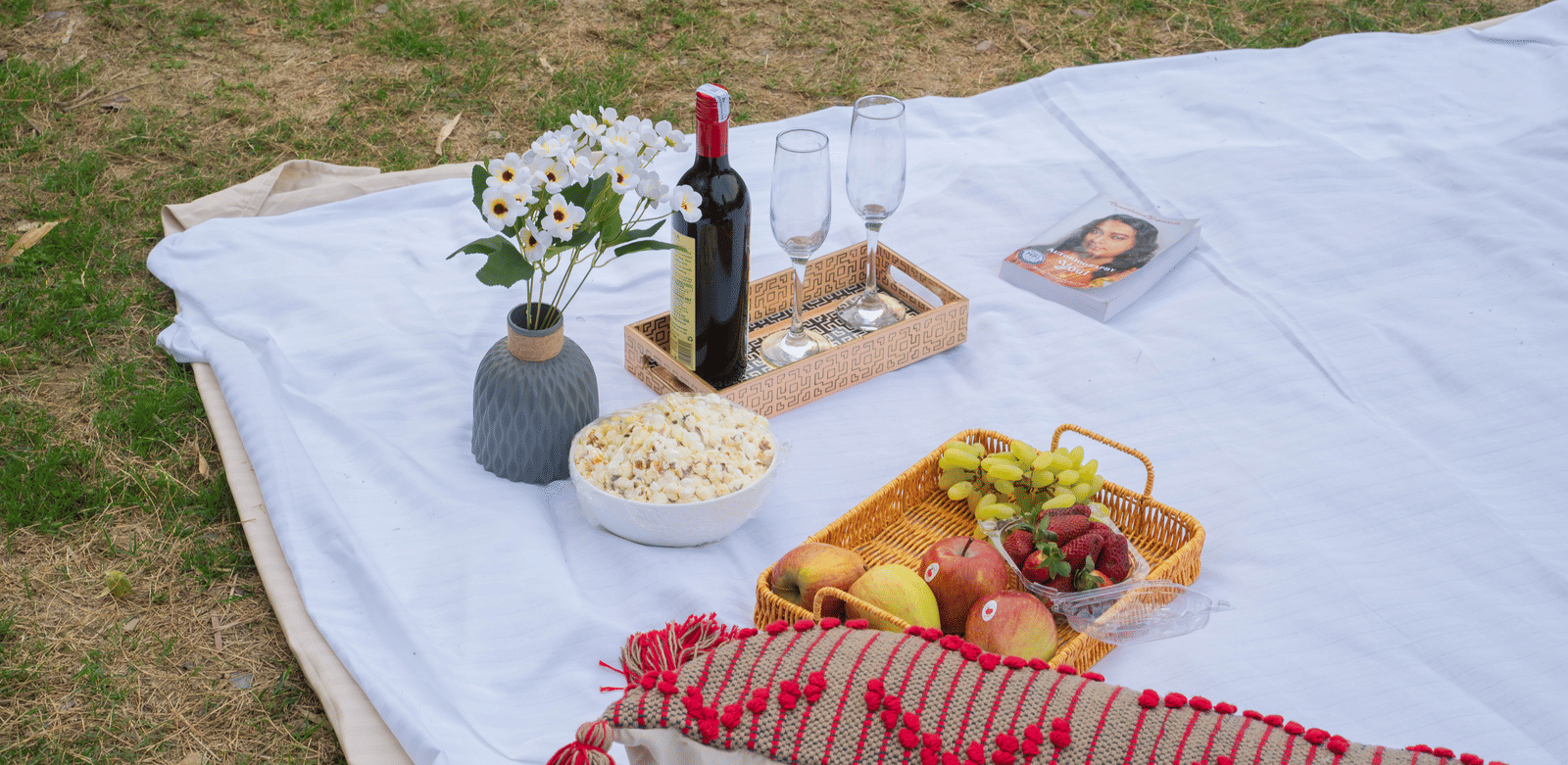 A picnic blanket on the grass is laid with fruit, wine, and cheese.