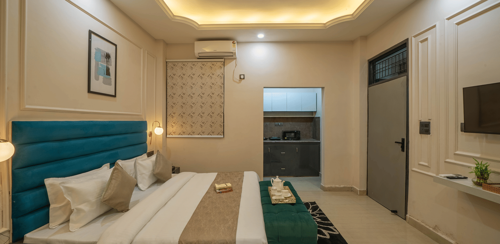 Spacious room with a bed, a teal tufted headboard, a white wardrobe, and a small kitchenette with dark grey lower cabinets next to the sleeping area - Perfectstayz Value Aalaya Suites, Haridwar