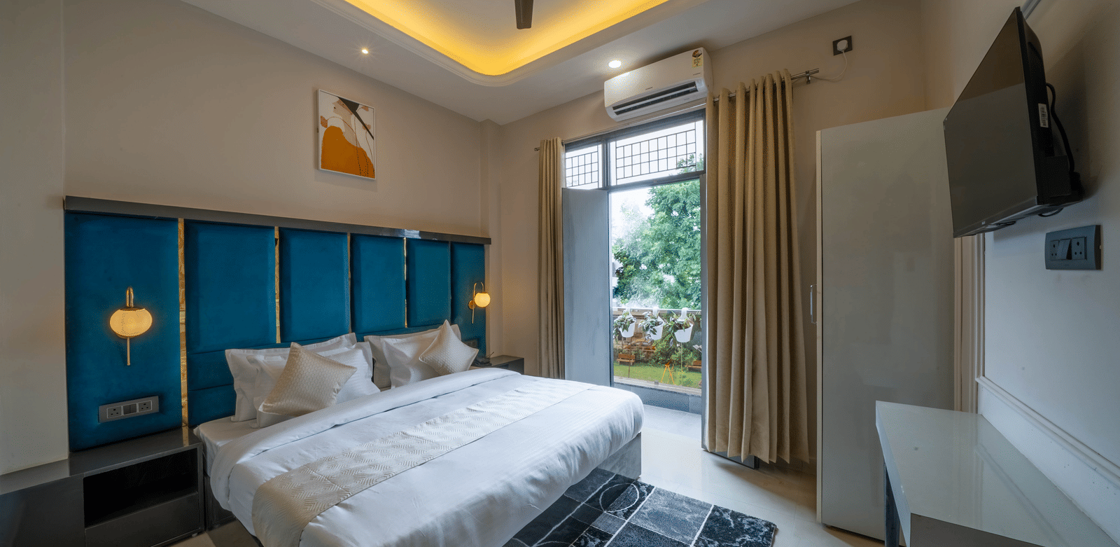 A spacious bedroom featuring a bed with a teal tufted headboard, large window with sheer curtains, a desk, a ceiling fan, and a television - Perfectstayz Value Aalaya Suites, Haridwar