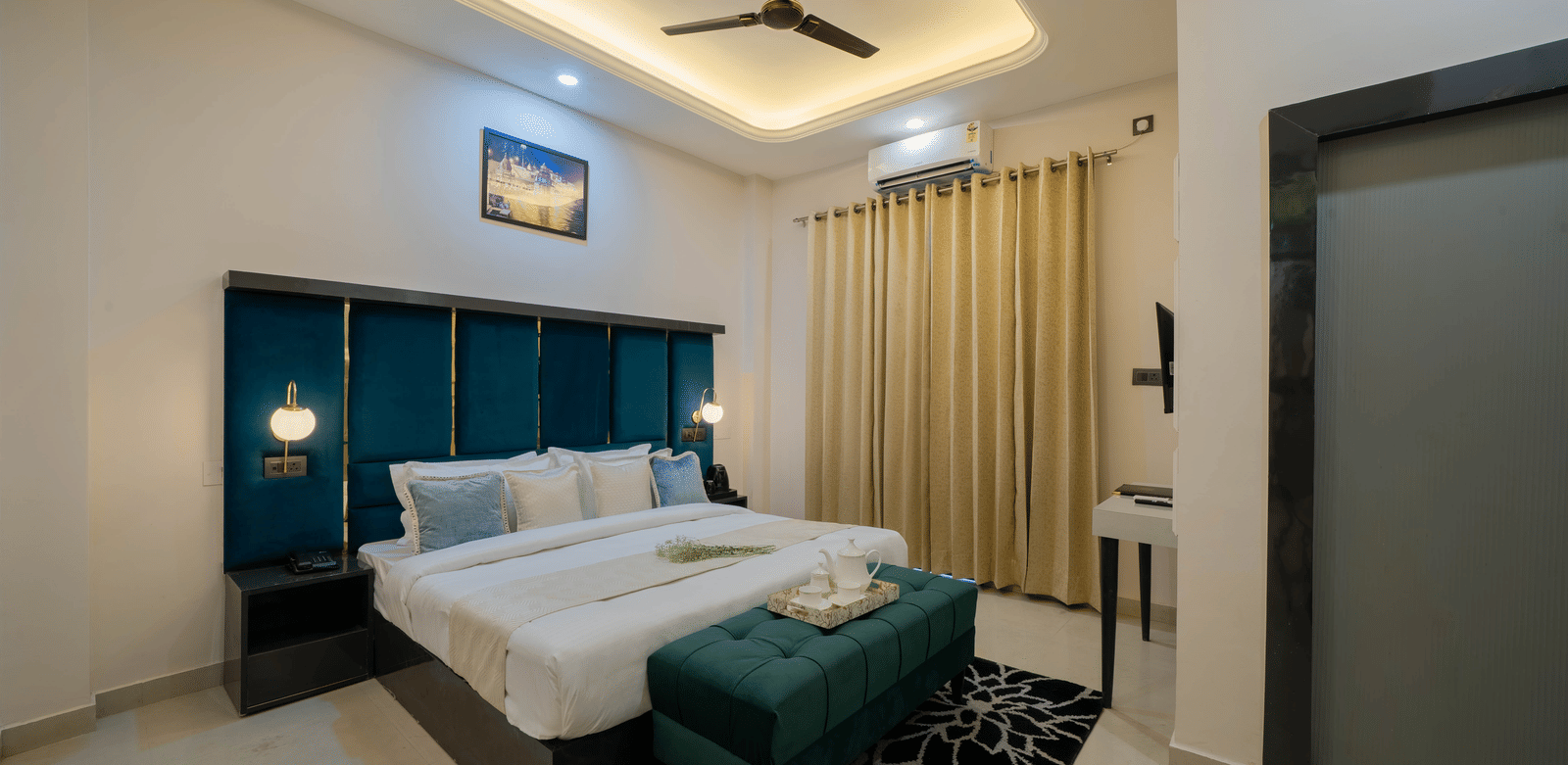 Interior view of a modern bedroom with a bed featuring a teal tufted headboard, white linens, a ceiling fan, and a dark green velvet bench - Perfectstayz Value Aalaya Suites, Haridwar